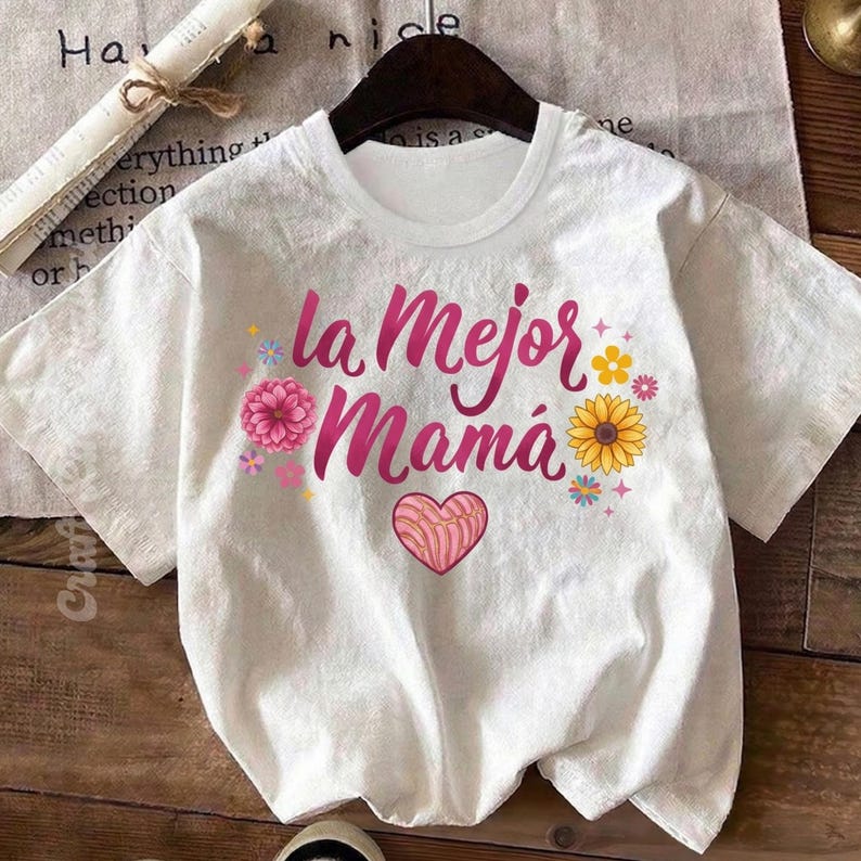 May include: White t-shirt with the text "la Mejor Mam&aacute;" in pink script, decorated with a pink flower, a sunflower, and a heart-shaped concha. The shirt is displayed on a wooden surface.