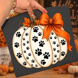 May include: A square canvas print featuring a white pumpkin decorated with black paw prints against a dark gray background. The pumpkin is topped with orange autumn leaves and a large, vibrant orange bow tied around its stem.
