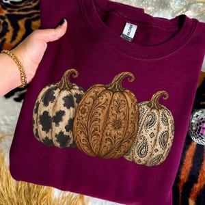 Pu&ograve; includere: Una felpa girocollo bordeaux con una grafica raffigurante tre zucche. La zucca a sinistra ha una stampa muccata bianca e nera, quella centrale un motivo floreale marrone e quella a destra una stampa paisley bianca e nera.