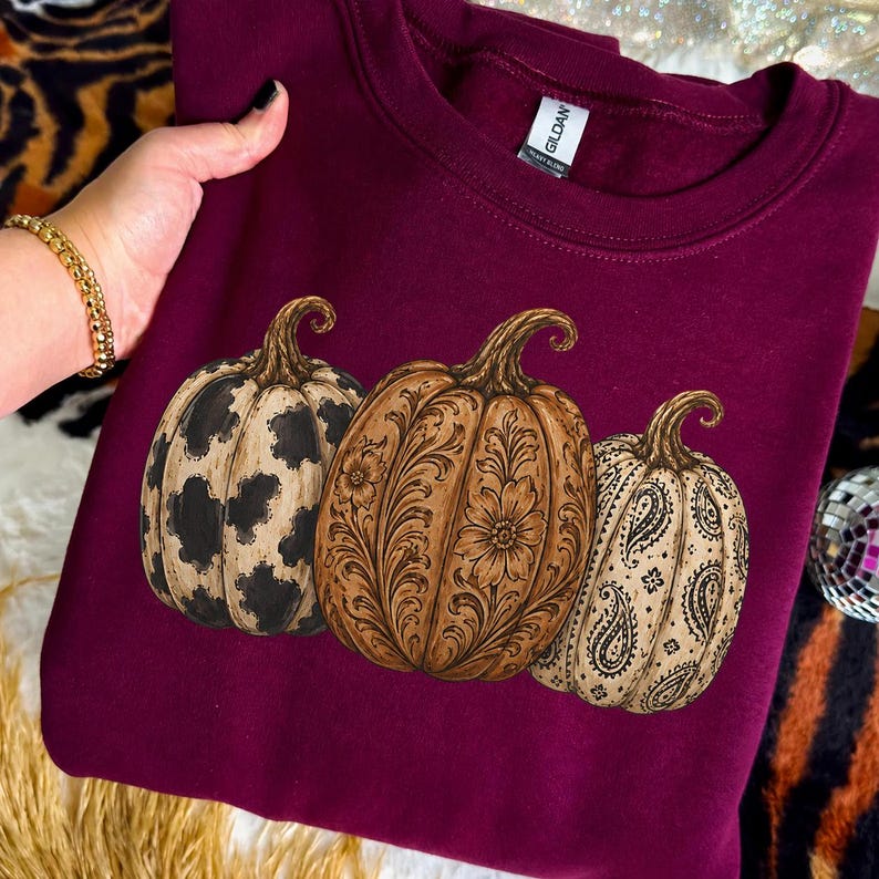Pu&ograve; includere: Una felpa girocollo bordeaux con una grafica raffigurante tre zucche. La zucca a sinistra ha una stampa muccata bianca e nera, quella centrale un motivo floreale marrone e quella a destra una stampa paisley bianca e nera.