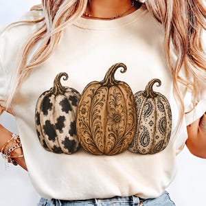 Pu&ograve; includere: Una t-shirt color crema con una grafica raffigurante tre zucche. La zucca a sinistra presenta una stampa muccata bianca e nera, quella centrale mostra un intricato motivo floreale marrone e quella a destra ha una stampa paisley nera.