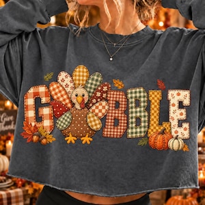 May include: A dark gray long-sleeve sweatshirt featuring a graphic of a turkey with a plaid-patterned body and tail feathers. The word GOBBLE is spelled out in large, blocky, plaid-patterned letters. The design includes orange leaves, acorns, and pumpkins.