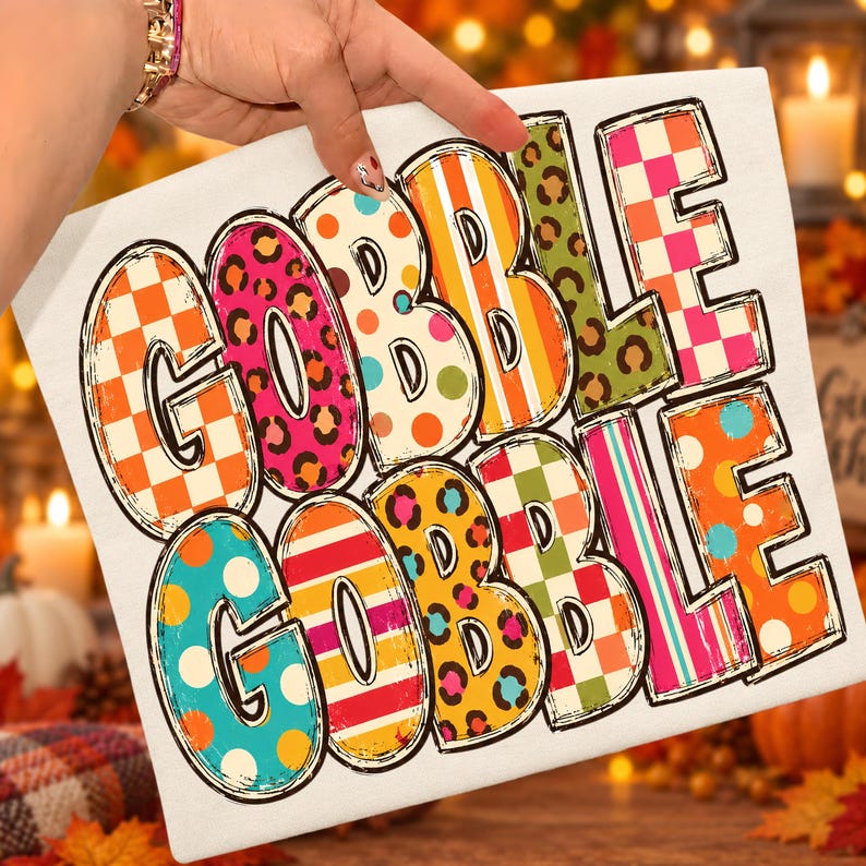 May include: A white fabric surface featuring the text GOBBLE GOBBLE in large, bold, block letters. Each letter is filled with a different colorful pattern, including orange and white checkers, pink leopard print, orange polka dots, and stripes.