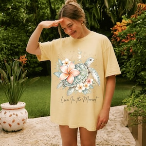 May include: A light yellow short-sleeve t-shirt featuring a graphic of a sea turtle surrounded by pink and white plumeria flowers and moon phase symbols. The text "Live In The Moment" is printed in black cursive script below the turtle illustration.