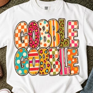May include: A white short-sleeve t-shirt featuring the text GOBBLE GOBBLE printed in two rows. Each letter is filled with a different colorful pattern, including orange and white checkers, pink leopard print, polka dots, stripes, and green leopard print.