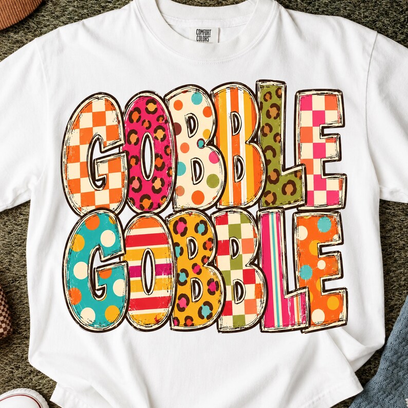 May include: A white short-sleeve t-shirt featuring the text GOBBLE GOBBLE printed in two rows. Each letter is filled with a different colorful pattern, including orange and white checkers, pink leopard print, polka dots, stripes, and green leopard print.