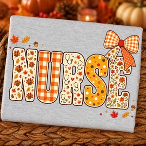 May include: A light gray sweatshirt featuring the word NURSE in large block letters. The letters are decorated with autumn-themed patterns including turkeys, stethoscopes, leaves, acorns, and polka dots, topped with a large orange plaid bow.