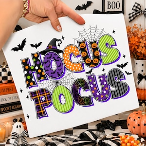 Könnte beinhalten: Ein weißes rechteckiges Schild mit dem Text HOCUS POCUS in großen, bunten, gemusterten Buchstaben. Das Design enthält orangefarbene Kürbisdrucke, violette Fledermausmuster, grüne Streifen und schwarze Spinnennetze, akzentuiert durch Fledermäuse.