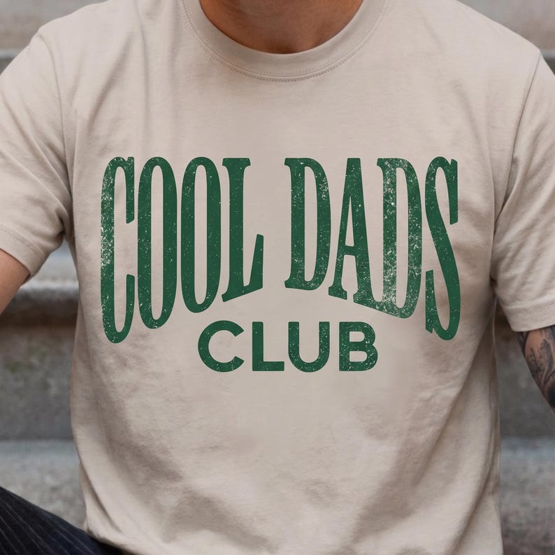 May include: A beige t-shirt with the words "COOL DADS CLUB" printed in a distressed green font. The text is centered on the shirt, with "COOL DADS" stacked above "CLUB". The shirt has short sleeves and a crew neck.