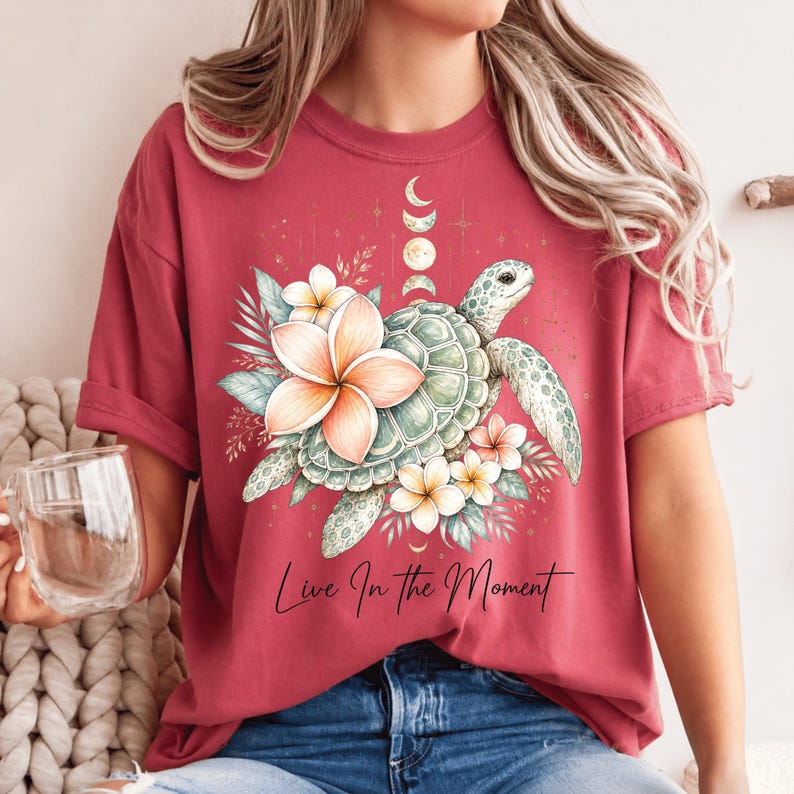 May include: A dark red short-sleeve t-shirt featuring a graphic of a sea turtle surrounded by tropical flowers and a moon phase cycle. The text "Live In The Moment" is printed in black script below the turtle and floral illustration.