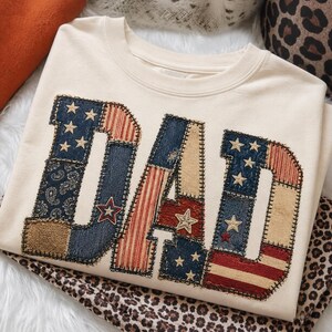 Könnte beinhalten: Cremefarbenes Sweatshirt mit dem Wort "DAD" in einem Patchwork-Design appliziert. Die Buchstaben weisen patriotische Muster auf, darunter Sterne, Streifen und Paisley, in Blau-, Rot- und Beigetönen. Ein Stern ist auf dem Buchstaben "A" aufgenäht.
