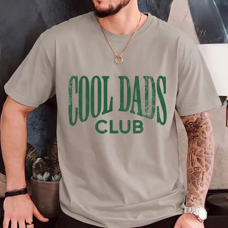 May include: A light gray t-shirt with the text "COOL DADS CLUB" in distressed green lettering. The shirt has short sleeves and a crew neck. The image shows the shirt on a person.