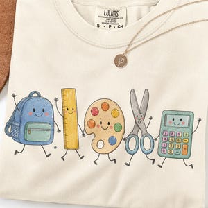 May include: Cream-colored t-shirt featuring a graphic of animated school supplies: a backpack, ruler, paint palette, scissors, and calculator. The shirt has the text "LULURS" on the upper part. A gold necklace with a pendant is also visible.