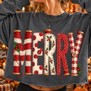 Könnte beinhalten: Ein anthrazitfarbenes Langarm-Sweatshirt mit dem Wort MERRY in großen, dekorativen Buchstaben. Jeder Buchstabe ist wie eine Garnspule gestaltet, mit rot-weißen Streifen, Punkten und Karomustern, verziert mit Stechpalmenzweigen.