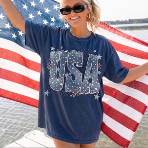 May include: Navy blue t-shirt with the word "USA" in a white and blue star-filled design. The shirt is worn with sunglasses and gold jewelry, and the person is holding an American flag.