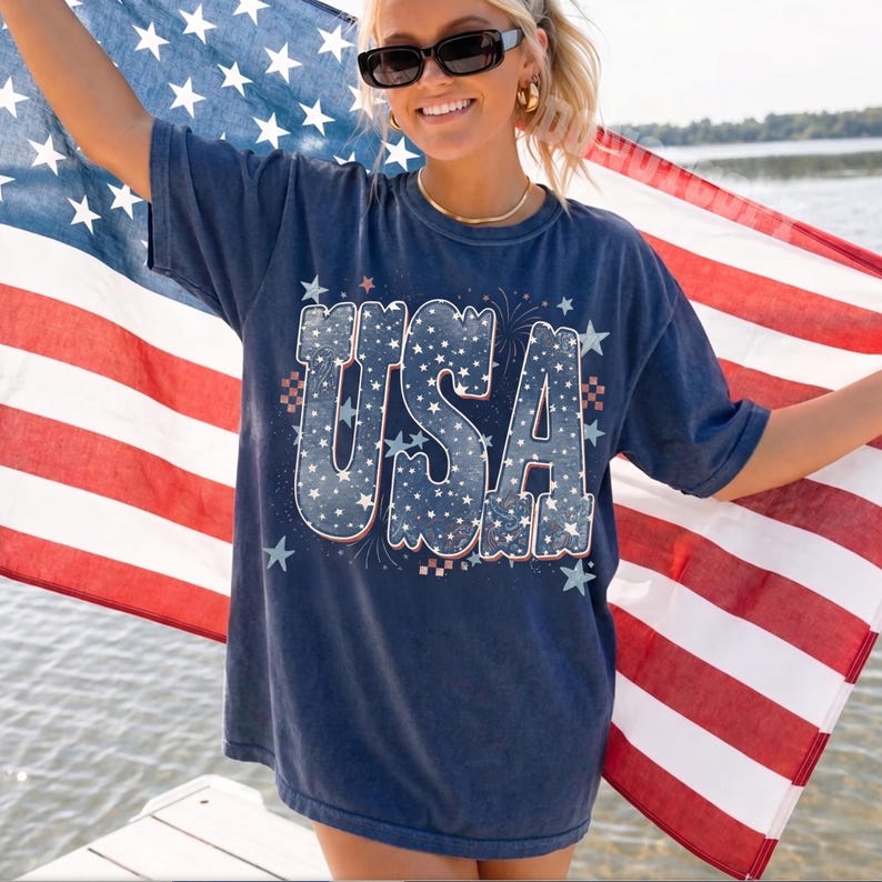 May include: Navy blue t-shirt with the word "USA" in a white and blue star-filled design. The shirt is worn with sunglasses and gold jewelry, and the person is holding an American flag.