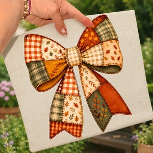 Könnte beinhalten: Eine quadratische Leinwand mit der Illustration einer großen, dekorativen Schleife, die aus verschiedenen Stoffmustern im Patchwork-Stil besteht. Das Design umfasst orange-weißes Vichy-Karo, dunkelgrünes Schottenkaro und cremefarbenen Stoff mit Herbstblattmotiven.