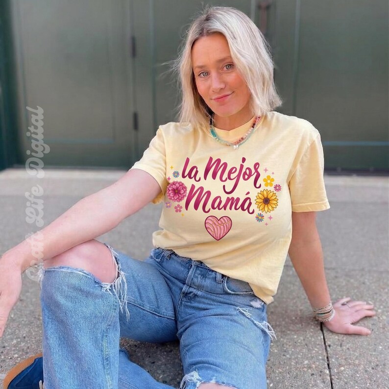 May include: A light yellow t-shirt with the text "la Mejor Mam&aacute;" in pink script, decorated with floral and heart graphics. The shirt is paired with blue jeans. The image shows a person wearing the shirt.