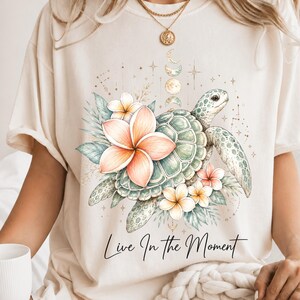 May include: A cream-colored t-shirt featuring a graphic of a sea turtle surrounded by pink and white plumeria flowers, green leaves, and a vertical sequence of moon phases. The text "Live In The Moment" is printed in black script below the design.
