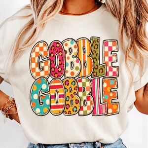 May include: A cream-colored crewneck sweatshirt featuring the text GOBBLE GOBBLE in large, bold letters. Each letter is filled with a different colorful pattern, including orange and white checkers, pink leopard print, polka dots, and stripes.