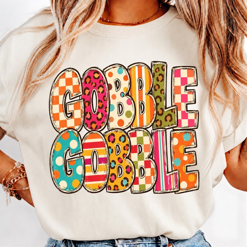 May include: A cream-colored crewneck sweatshirt featuring the text GOBBLE GOBBLE in large, bold letters. Each letter is filled with a different colorful pattern, including orange and white checkers, pink leopard print, polka dots, and stripes.