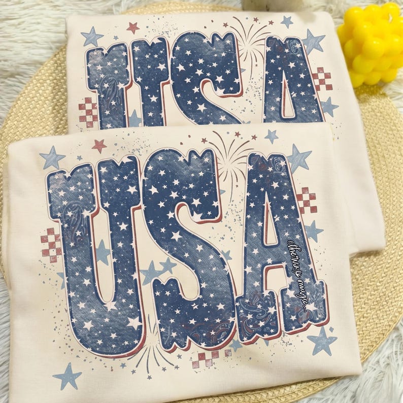 May include: Two cream-colored t-shirts with the word "USA" in large, distressed blue letters filled with white stars. Red and blue accents include fireworks, stars, and checkered patterns. The text "there is magic" is printed on the bottom right of the shirt.