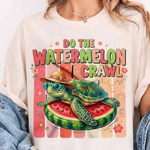 May include: A cream-colored t-shirt featuring a cartoon turtle wearing a hat and glasses, lounging on a watermelon slice. The text "DO THE WATERMELON CRAWL" is above the turtle in colorful letters. The design includes floral and star accents.
