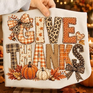 May include: A white t-shirt featuring the text GIVE THANKS in large, patterned block letters. The letters include plaid, leopard print, and floral designs. The graphic is decorated with two pumpkins, autumn leaves, acorns, and two bows.