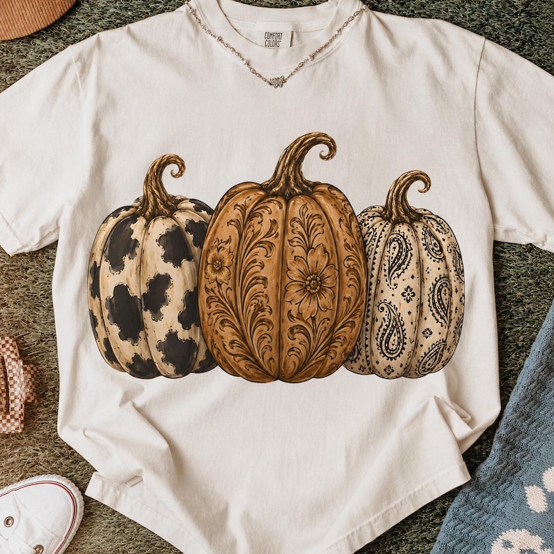 Pu&ograve; includere: Una t-shirt bianca a maniche corte con una grafica raffigurante tre zucche. La zucca a sinistra ha una stampa muccata bianca e nera, quella centrale presenta un intricato motivo floreale marrone e quella a destra ha una stampa paisley nera.