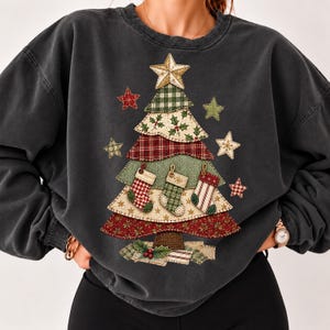 Könnte beinhalten: Ein anthrazitfarbenes Sweatshirt mit Rundhalsausschnitt und einem großen Grafikdruck eines stilisierten Weihnachtsbaums aus verschiedenen Stoffmustern. Der Baum ist mit drei hängenden Strümpfen, einem Stern als Spitze und kleinen Sternen verziert.