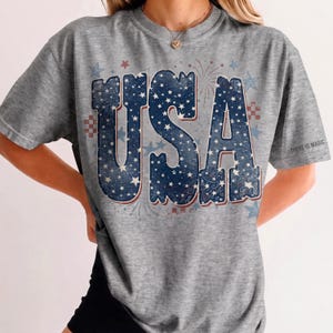 May include: Gray t-shirt with a patriotic "USA" design in blue and white, featuring stars and fireworks. The text "THERE IS MAGIC" is printed on the sleeve. A casual, comfortable tee for celebrating American pride.