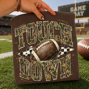 May include: A dark brown t-shirt featuring the text TOUCH DOWN in large block letters with a camouflage pattern. A brown football with white laces is centered between the words, accompanied by a black and white checkered graphic element.