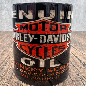 May include: Black ceramic mug with the Harley-Davidson logo in white and orange. The mug features the words "GENUINE MOTOR HARLEY-DAVIDSON CYCLES OIL". The cylindrical mug has a glossy finish.