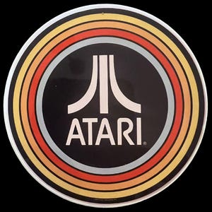 May include: A circular sign featuring the white Atari logo and text centred on a black background. The design is surrounded by concentric rings in shades of yellow, orange, and light blue, creating a retro aesthetic on a smooth, flat surface.