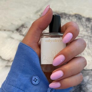 May include: A clear glass bottle with a black cap, labelled "oilyolive" with a floral design, held by a hand with pink nail polish. The person is wearing a blue button-down shirt.