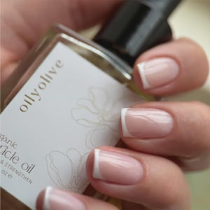 May include: A hand holds a bottle of "oilyolive" cuticle oil. The nails are painted with a French manicure, featuring white tips. The bottle has a white label with gold text and a floral design. The image focuses on nail care.