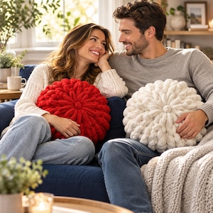May include: Two round, chunky knit pillows, one in vibrant red and one in cream white, featuring a textured, circular pattern with a central button-like knot. These decorative cushions are soft, thick, and have a distinct, hand-woven appearance.