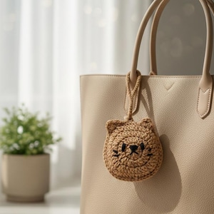 May include: A beige tote bag with a crocheted cat face charm. The charm, with black embroidered features, hangs from the handle. The bag is set against a soft, neutral background. The bag is made of a leather-like material.