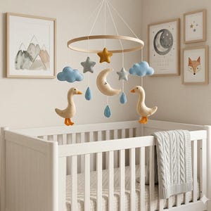 Puede incluir: Una cuna blanca con un móvil colgante con nubes de fieltro, estrellas, una luna y dos gansos. El móvil está suspendido sobre la cuna. Obras de arte de montañas, una luna y un zorro están en la pared.