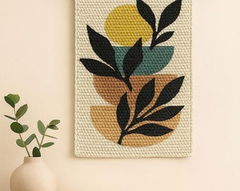 May include: A beige, rectangular, woven wall hanging with a minimalist design. It features a yellow circle, teal and brown semi-circles, and black leaf silhouettes. The artwork is suspended from a wooden dowel with a cream-colored string.