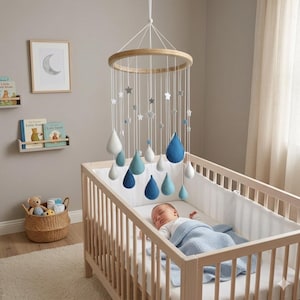 May include: A baby's nursery with a wooden crib and a hanging mobile. The mobile features a wooden ring with hanging stars and teardrop shapes in shades of blue and white. A baby sleeps peacefully in the crib, covered with a blue blanket.