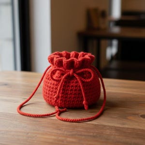 Może przedstawiać: Mała, okrągła, czerwona szydełkowa torba ze sznurkiem i pasującym zamknięciem na sznurek. Torba leży na drewnianej powierzchni. Torba ma prosty, teksturowany design i swobodną estetykę.