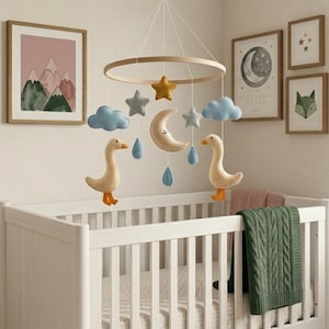 May include: A nursery scene featuring a white crib with a hanging mobile. The mobile has felt geese, clouds, stars, a moon, and raindrops. Artwork on the wall includes mountain and celestial-themed framed prints. A green blanket is draped over the crib.