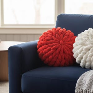 May include: A navy blue armchair holds two round, knitted pillows, one red and one white. A gray blanket is draped over the arm. A wooden coffee table with a plant and mug is in the background. The pillows add a cozy touch to the room.