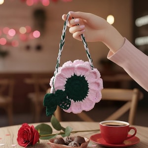 May include: A handmade pink and green crochet flower bag with a braided strap. The bag is held above a table with a red rose, chocolates, and a cup of coffee. The background has heart-shaped decorations.