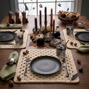 May include: A dining table setting featuring cream-colored crocheted placemats, matte black plates, and sage green cloth napkins with bat-shaped napkin rings. The centerpiece includes taper candles, small pumpkins, a skull, and a bowl of gourds.