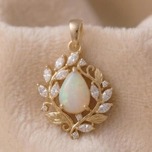 May include: A gold pendant featuring a pear-shaped opal gemstone at the center, surrounded by a wreath of leaves and marquise-cut diamonds. The pendant is suspended from a gold bail. The jewelry is set against a soft, beige background.