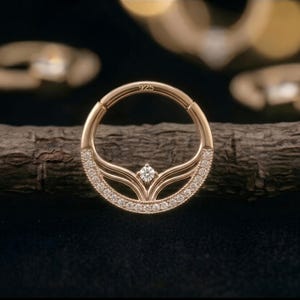 Könnte beinhalten: Ein goldfarbener Septumring mit Scharnierdesign. Der Ring ist mit kleinen, funkelnden Steinen am unteren Rand und einem einzelnen, größeren Stein in der Mitte verziert. Der Ring ist mit "925" markiert. Der Schmuck wird auf einem Holzstück präsentiert.