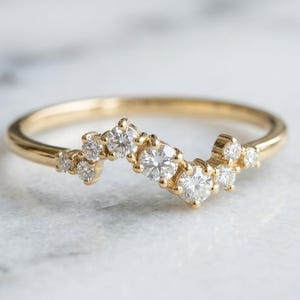 May include: A gold ring featuring a curved, wave-like band set with a cluster of nine round, clear, faceted stones of varying sizes arranged in a staggered, asymmetrical pattern across the top of the band, creating a delicate and sparkling appearance.