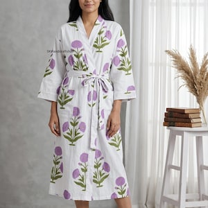 Pode incluir: Um roupão branco com um padrão floral repetitivo de flores circulares roxas e caules e folhas verdes. O roupão tem mangas compridas, decote em V e um cinto de tecido a condizer na cintura. O texto SKHandicraftshub é visível.